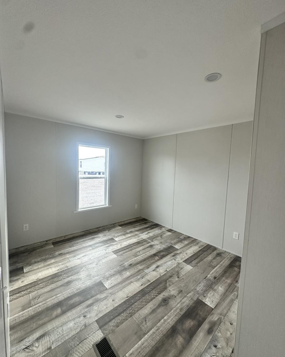 Empty room with light gray walls and wood-patterned flooring. A single window lets in natural light, creating a clean, minimalist atmosphere.