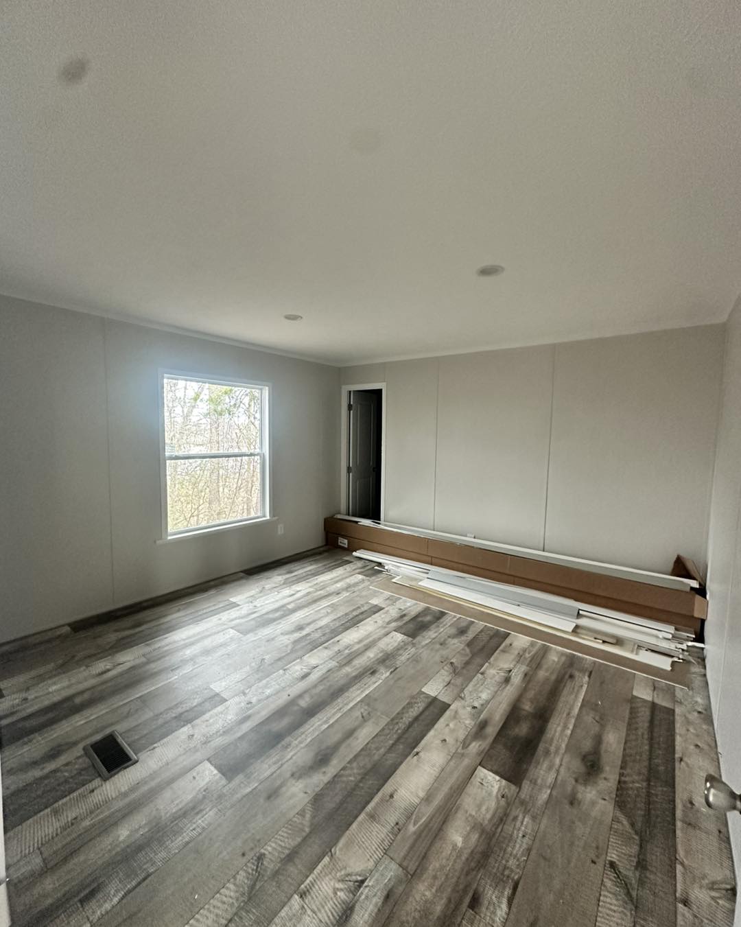 Empty room with gray wood flooring and pale walls, featuring a large window on the left. Stacked molding and materials lie against the far wall. Bright and minimal.