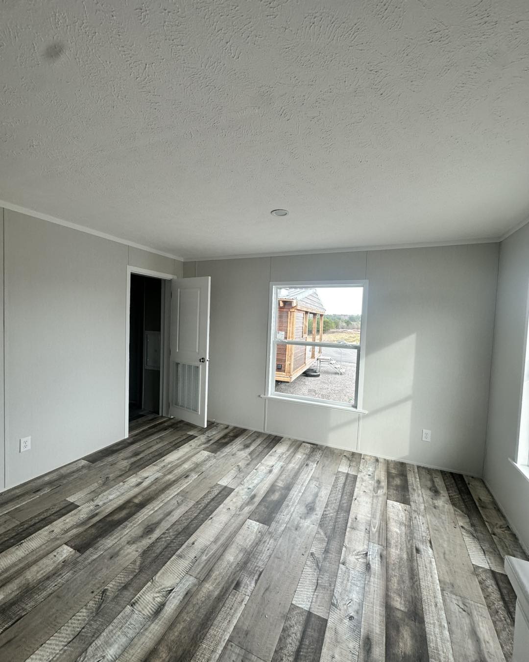 Empty room with gray walls and wood-textured vinyl flooring. A window shows a view of a small wooden structure outside. A white door is ajar.