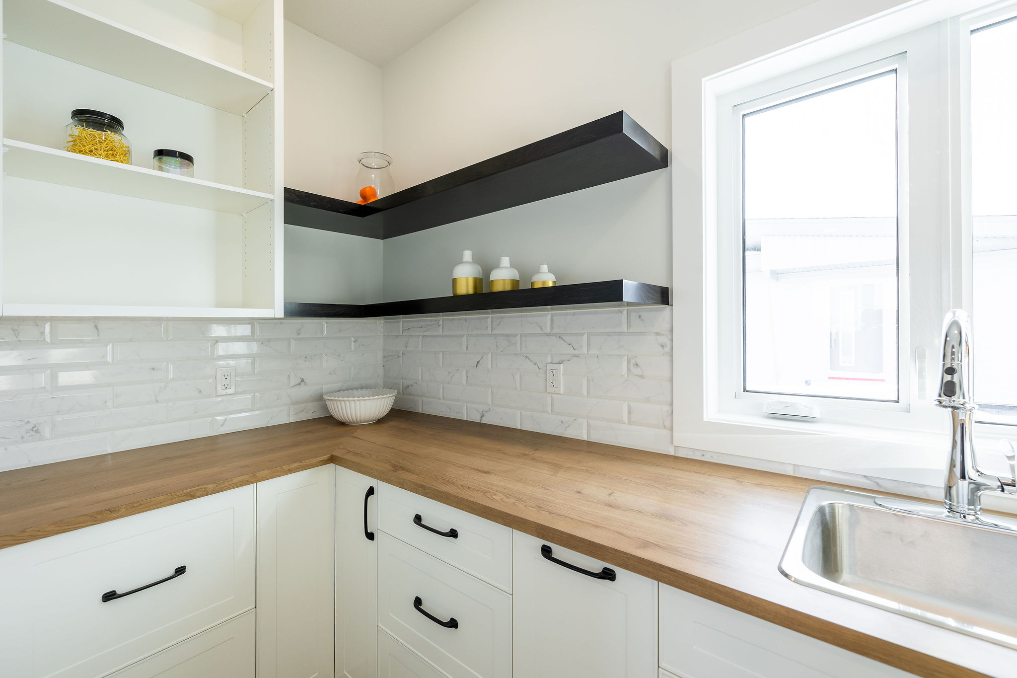 A modern kitchen corner with white cabinets, black handles, and wooden countertops. Open shelves hold jars and bottles. A sink is below a bright window.