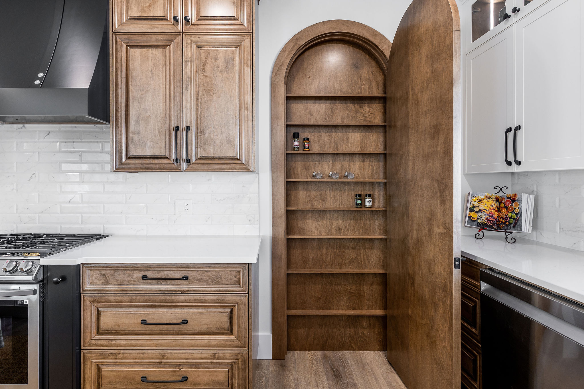 Kitchen with wooden cabinetry, arched hidden pantry door disguised as a bookshelf, white counters, and modern appliances. Cozy and functional design.