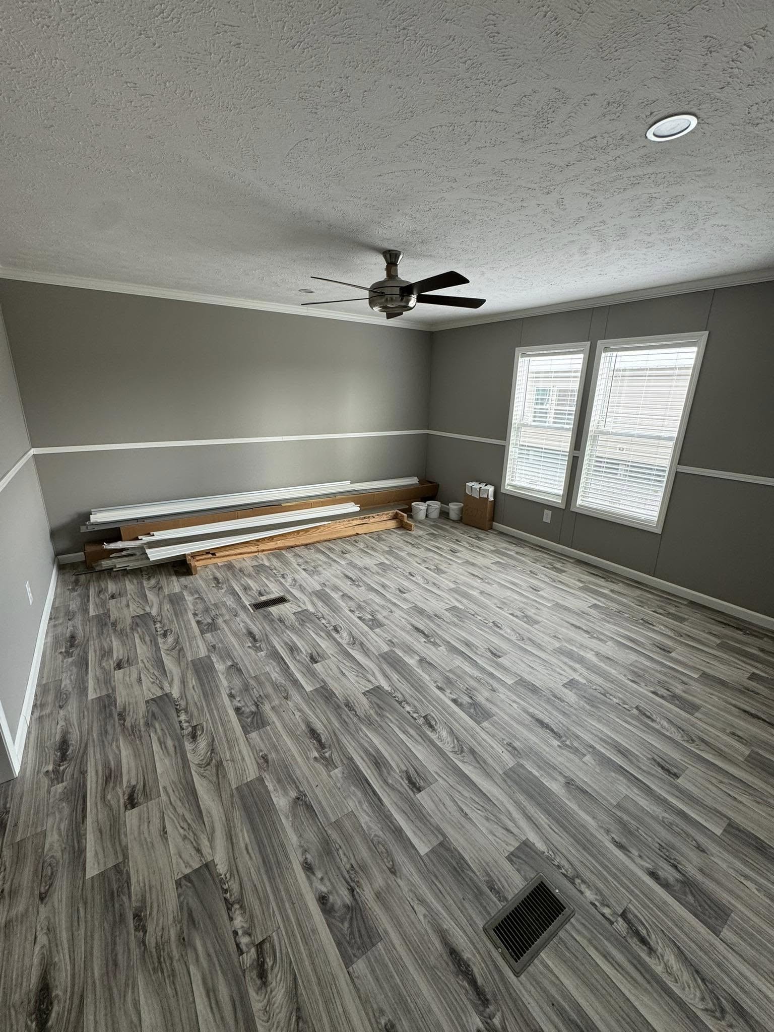 An empty room with gray laminate flooring and gray walls, featuring a ceiling fan. Windows let in natural light. Wood planks and paint supplies line the wall.