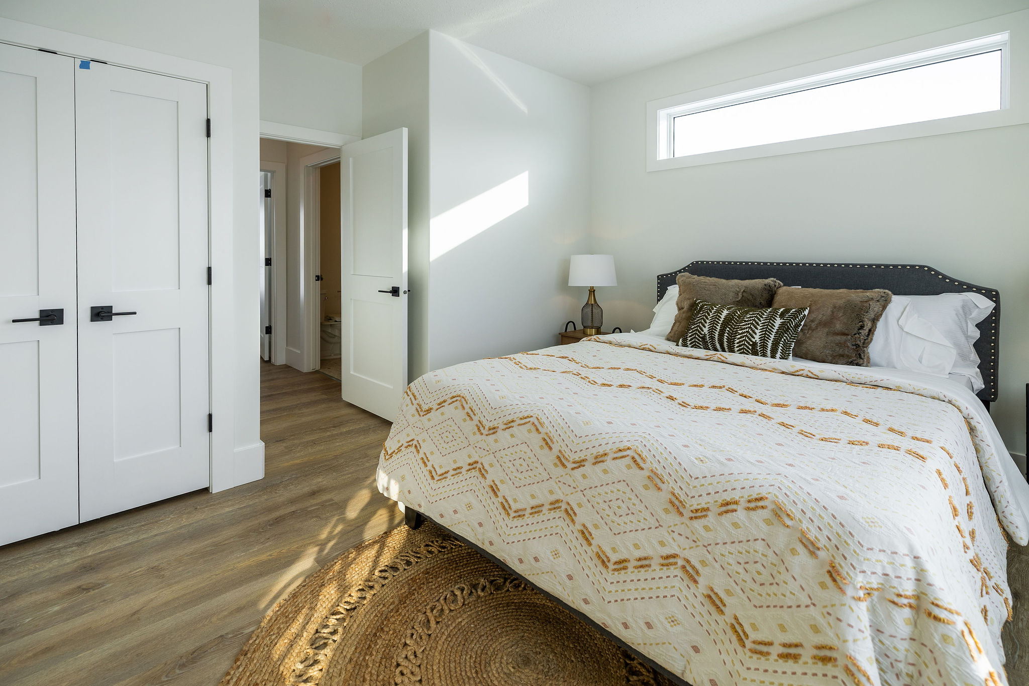A bright bedroom with light wooden floors, a neatly made bed with patterned bedding, decorative pillows, and a round woven rug. A lamp is on the bedside table by the bed with a window above. The overall ambience feels cozy and inviting.