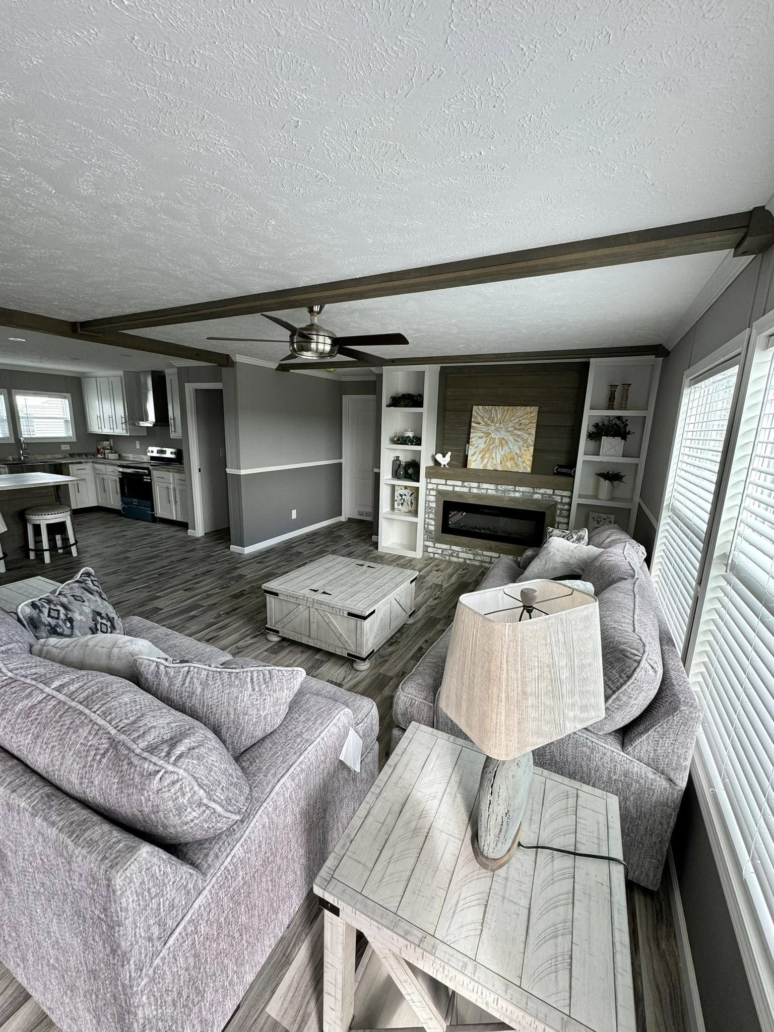 Modern living room with gray sofas, a wooden coffee table, and a fireplace. Ceiling fan overhead. Kitchen visible in the background. Cozy and inviting ambiance.
