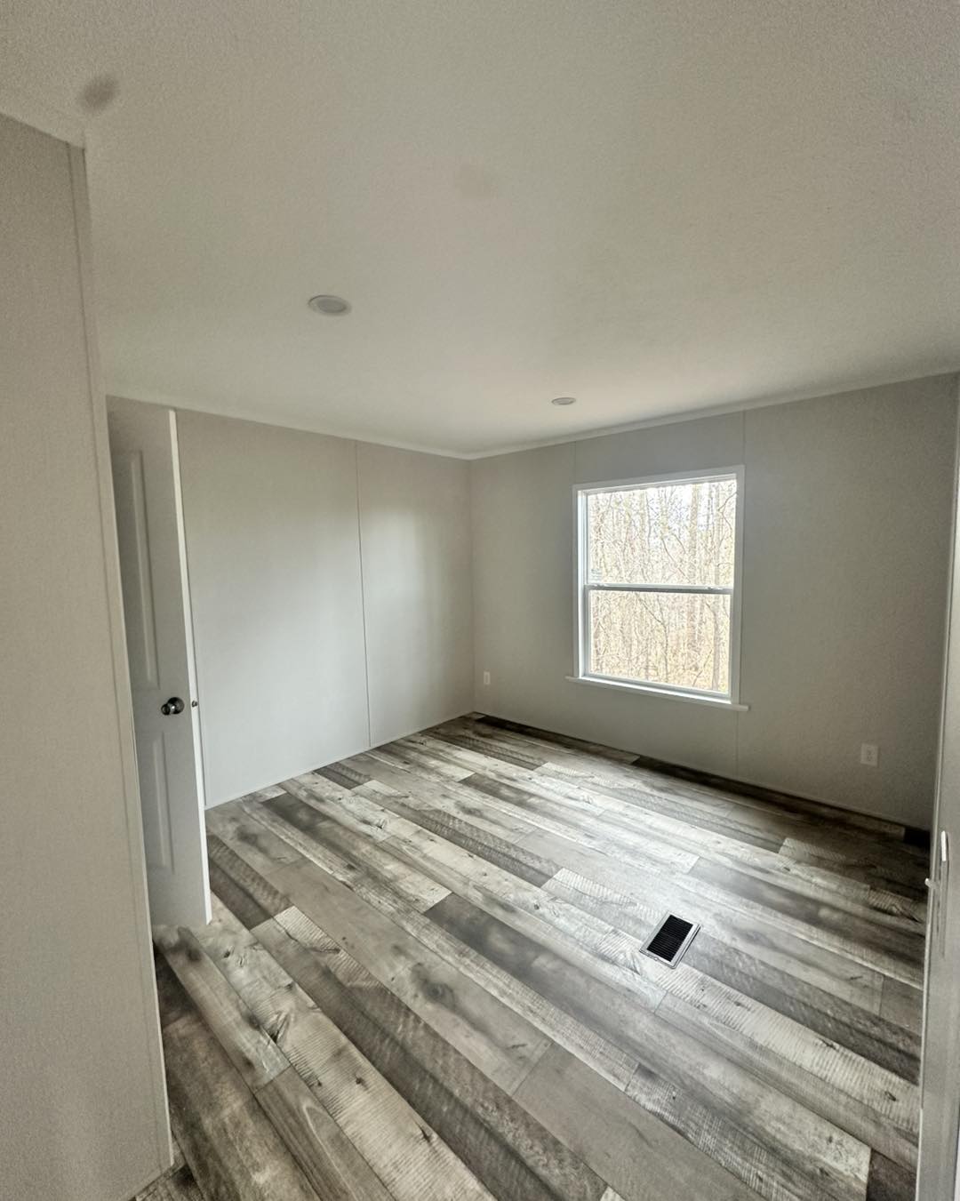 Empty room with light gray walls, dark wood laminate flooring, and a large window revealing tree branches. The space feels clean and spacious.