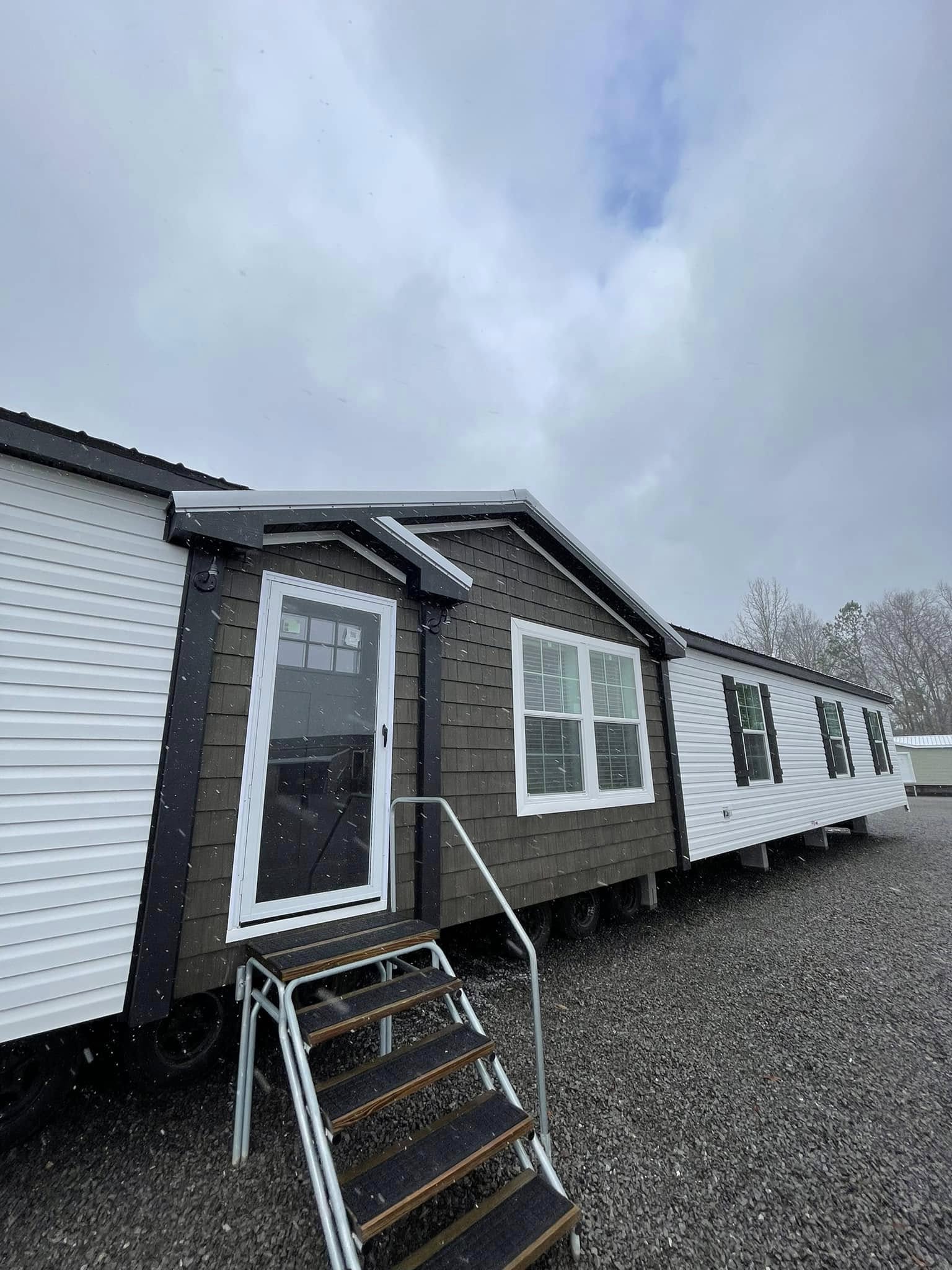 Mobile home with brown and white siding, front door and steps. Gravel ground and cloudy sky create a neutral, peaceful outdoor setting.