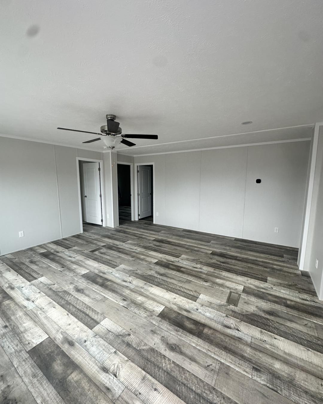 Spacious, empty room with light gray walls and rustic wood flooring. A ceiling fan is centered on the ceiling. Open doors lead to other rooms. Bright and modern feel.