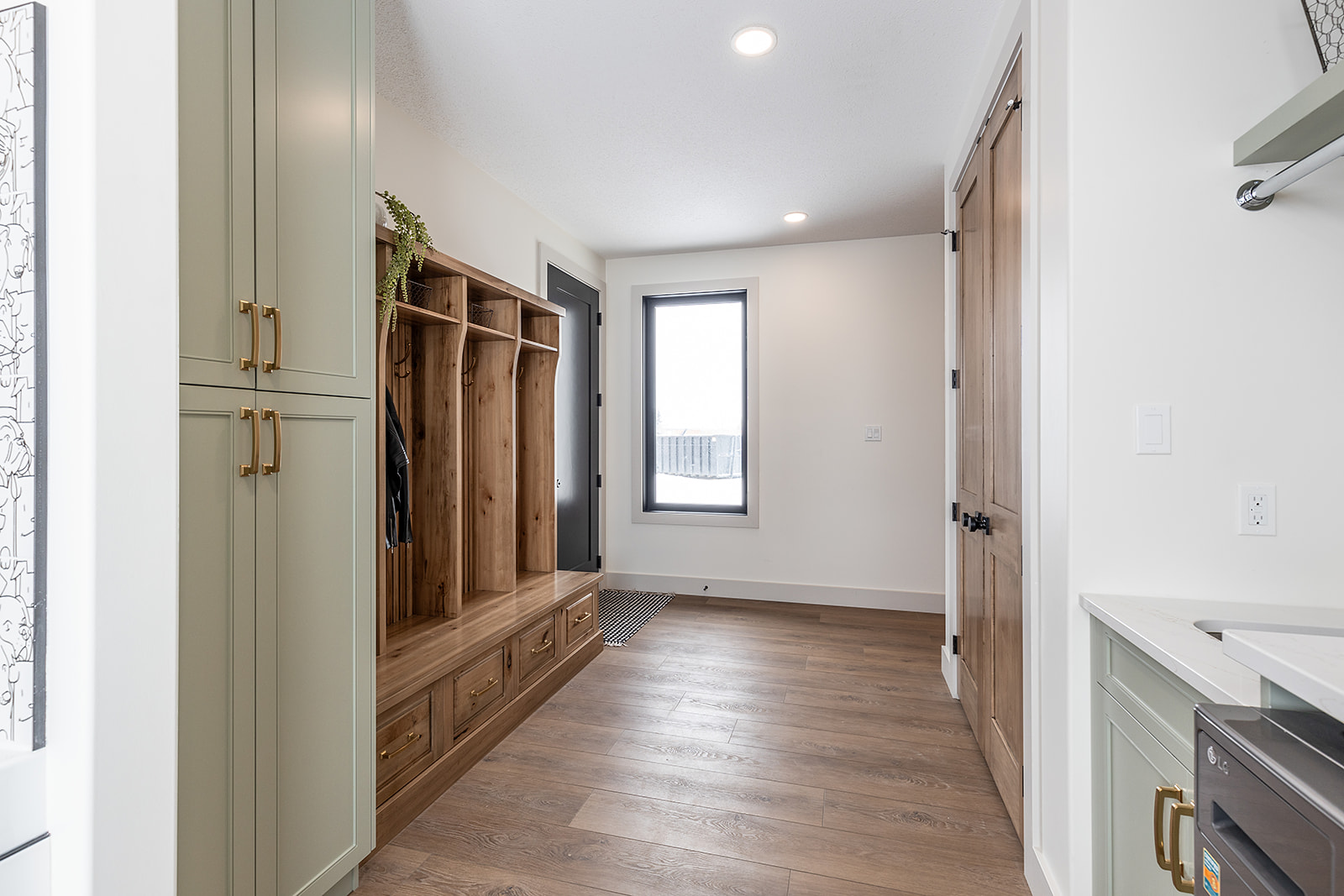 Bright mudroom with wooden lockers and drawers, green cabinets with brass handles, and a window. Calm, organized atmosphere.