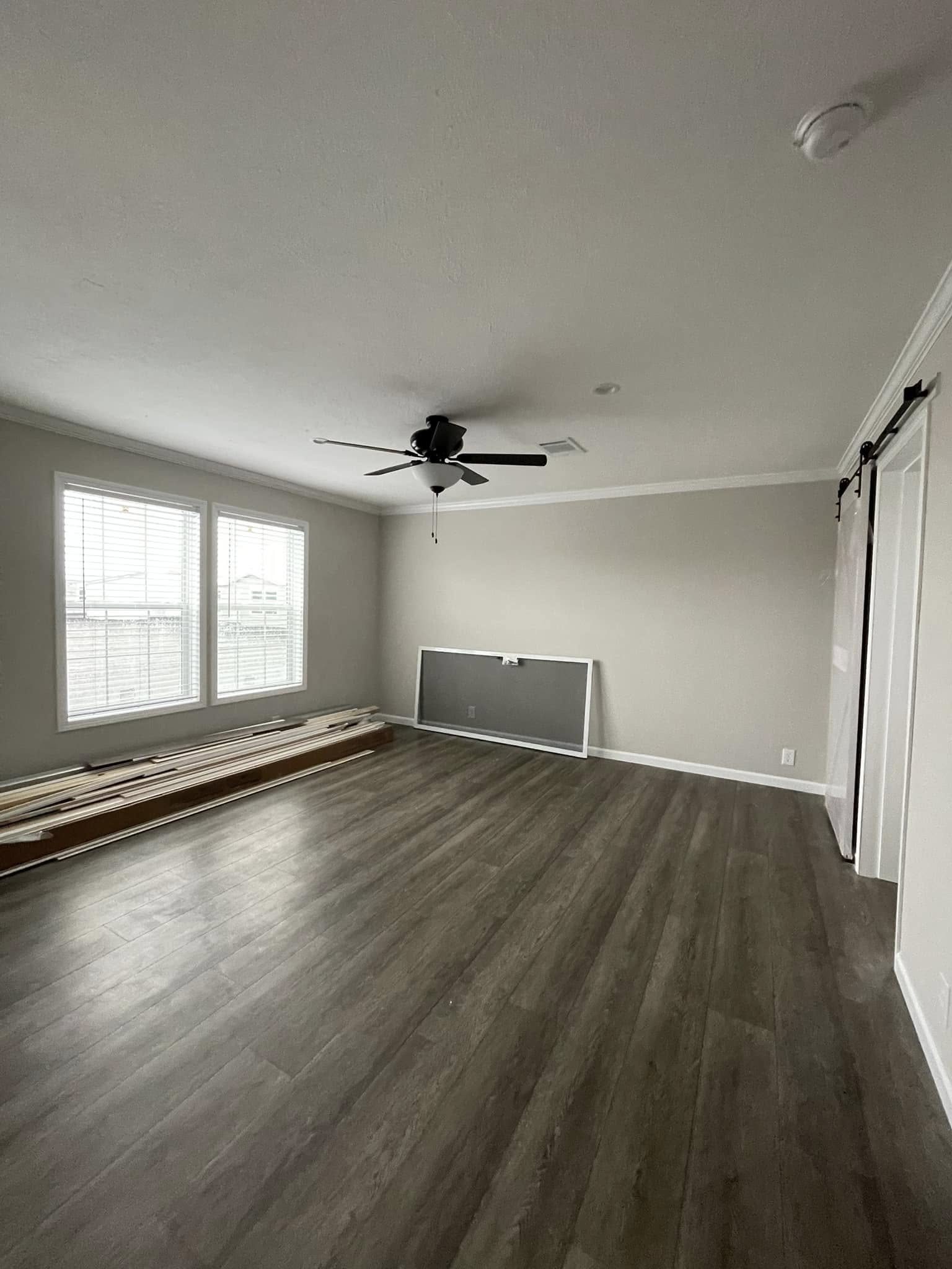 Spacious, empty room with light gray walls and wood flooring, featuring a ceiling fan, large windows, stacked wood planks, and a leaning screen door.