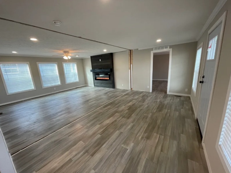 Spacious living room with light wood flooring, gray walls, and large windows. A modern electric fireplace adds a cozy touch. Bright, inviting ambiance.