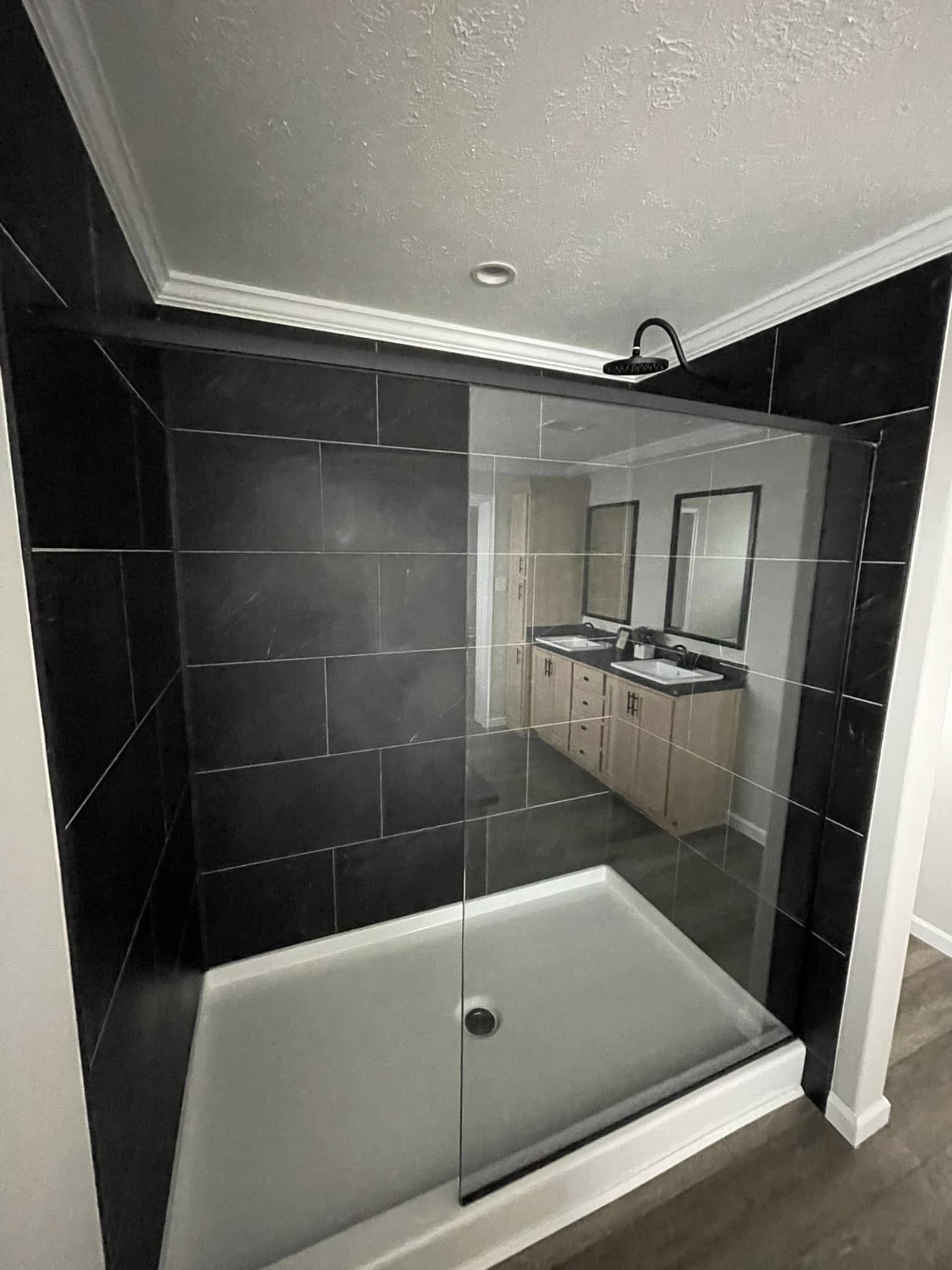 A modern bathroom features a spacious walk-in shower with dark tiles and glass doors, reflecting a double vanity with wood cabinets and dual mirrors.