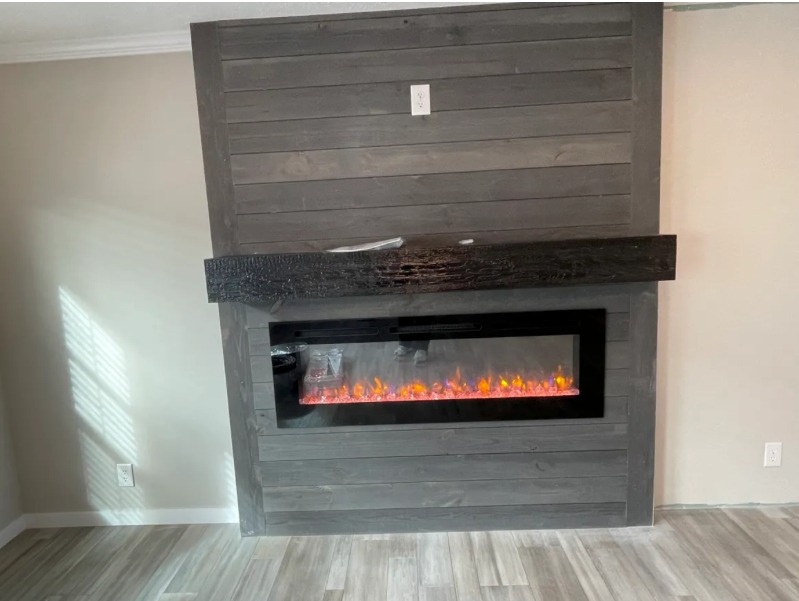 Dark gray shiplap wall featuring a modern electric fireplace with glowing orange flames. A chunky, charred wood mantel adds a rustic touch.