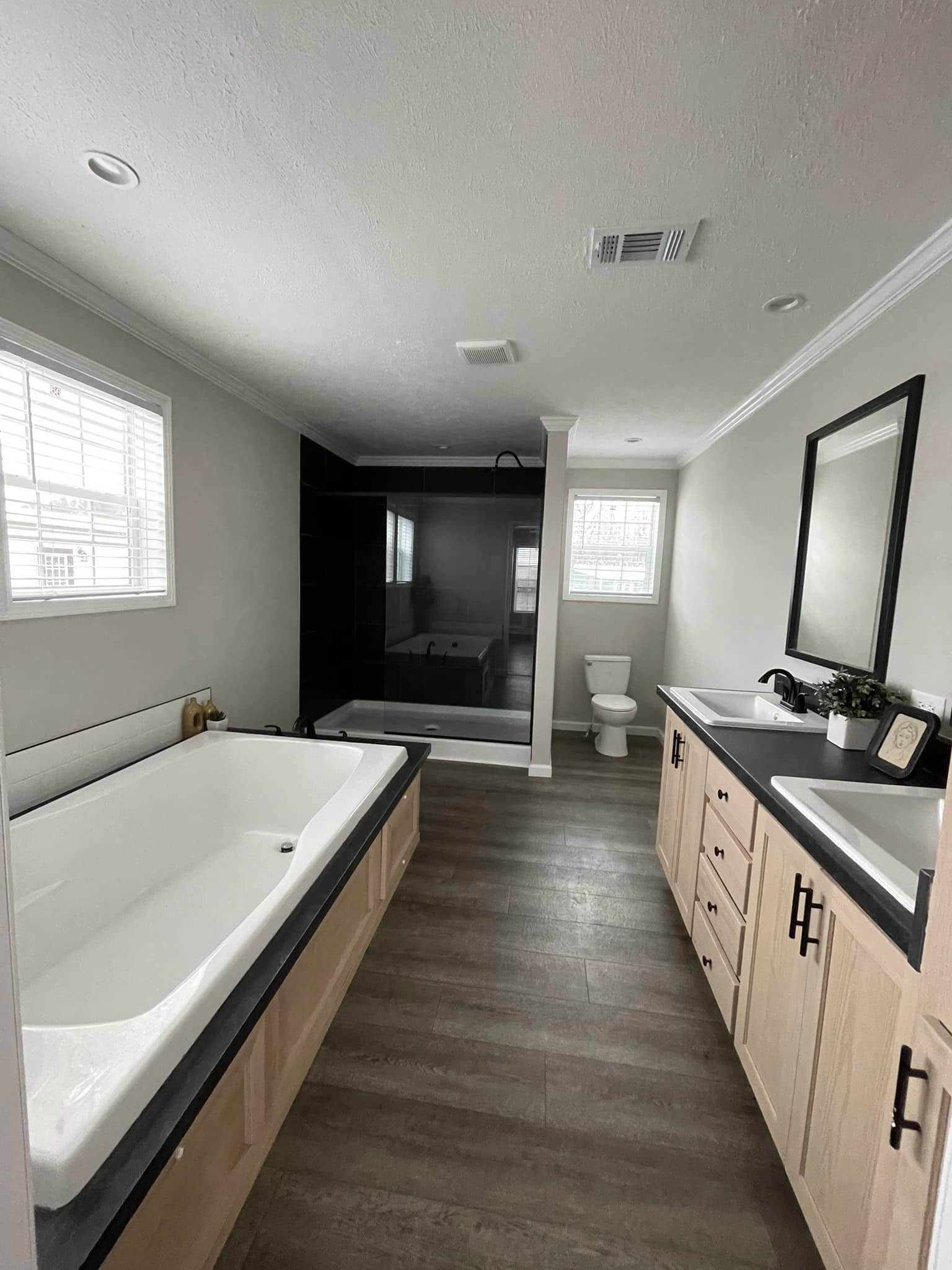 Modern bathroom with a large bathtub, black-tiled shower, wooden cabinets, and dual sinks. Bright and spacious with natural light from windows.