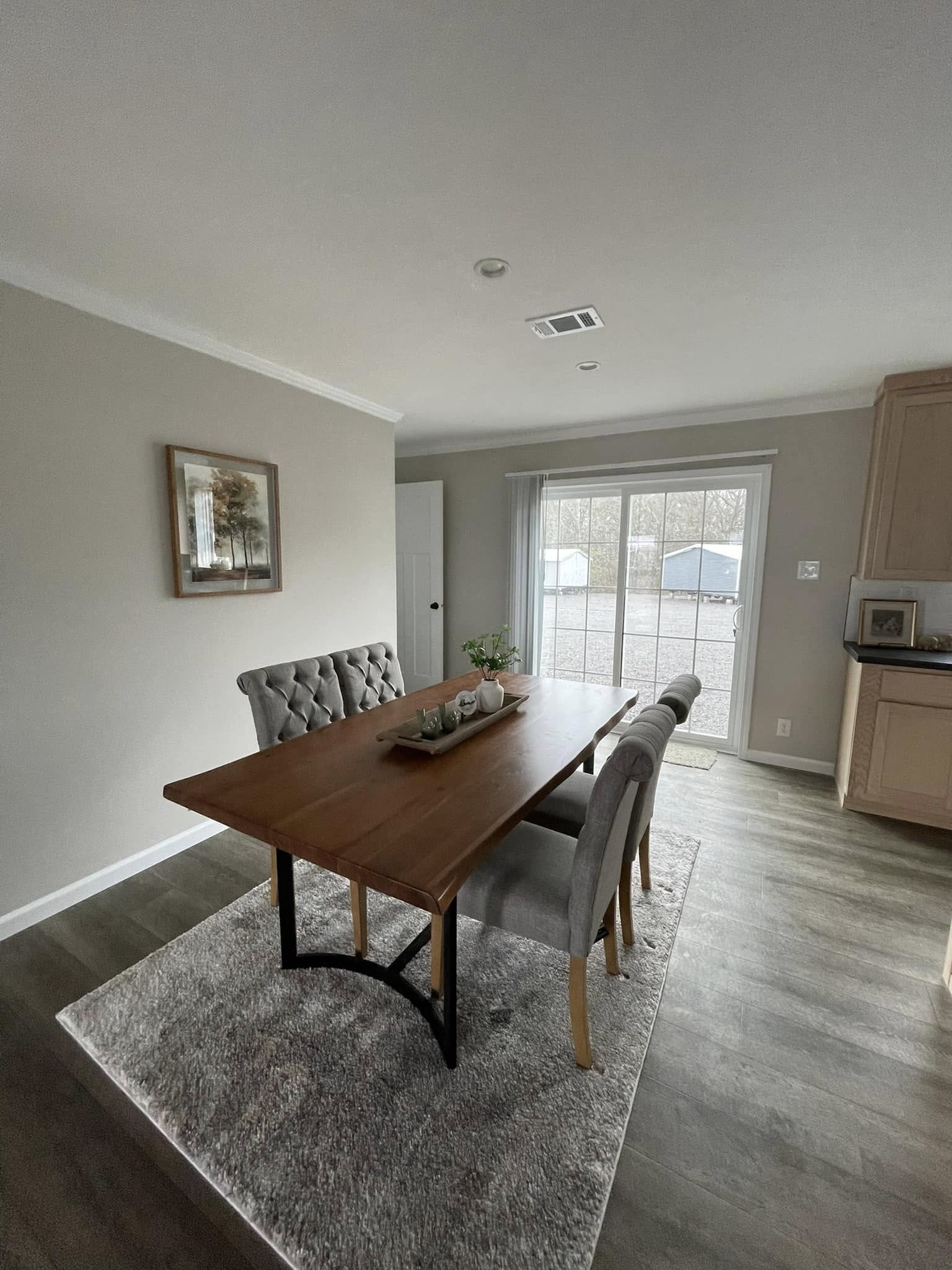 Bright dining room with a wooden table and four tufted chairs on a gray rug. Sliding glass doors offer a view of the outside, creating a cozy ambiance.