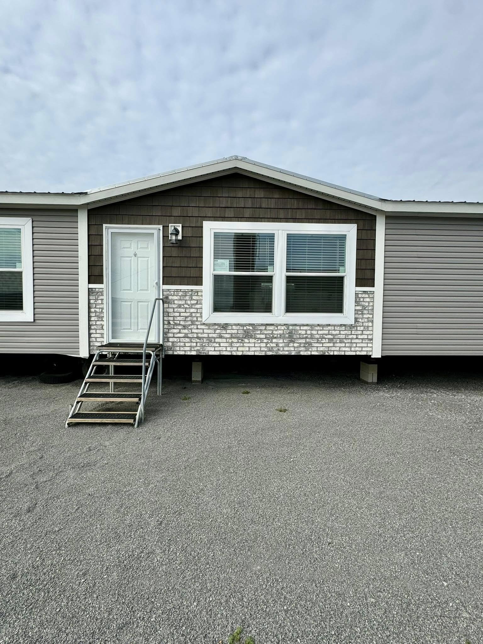 Single-story mobile home with gray siding and a pitched roof. It features a white door with three steps leading up. The mood is calm and minimalist.