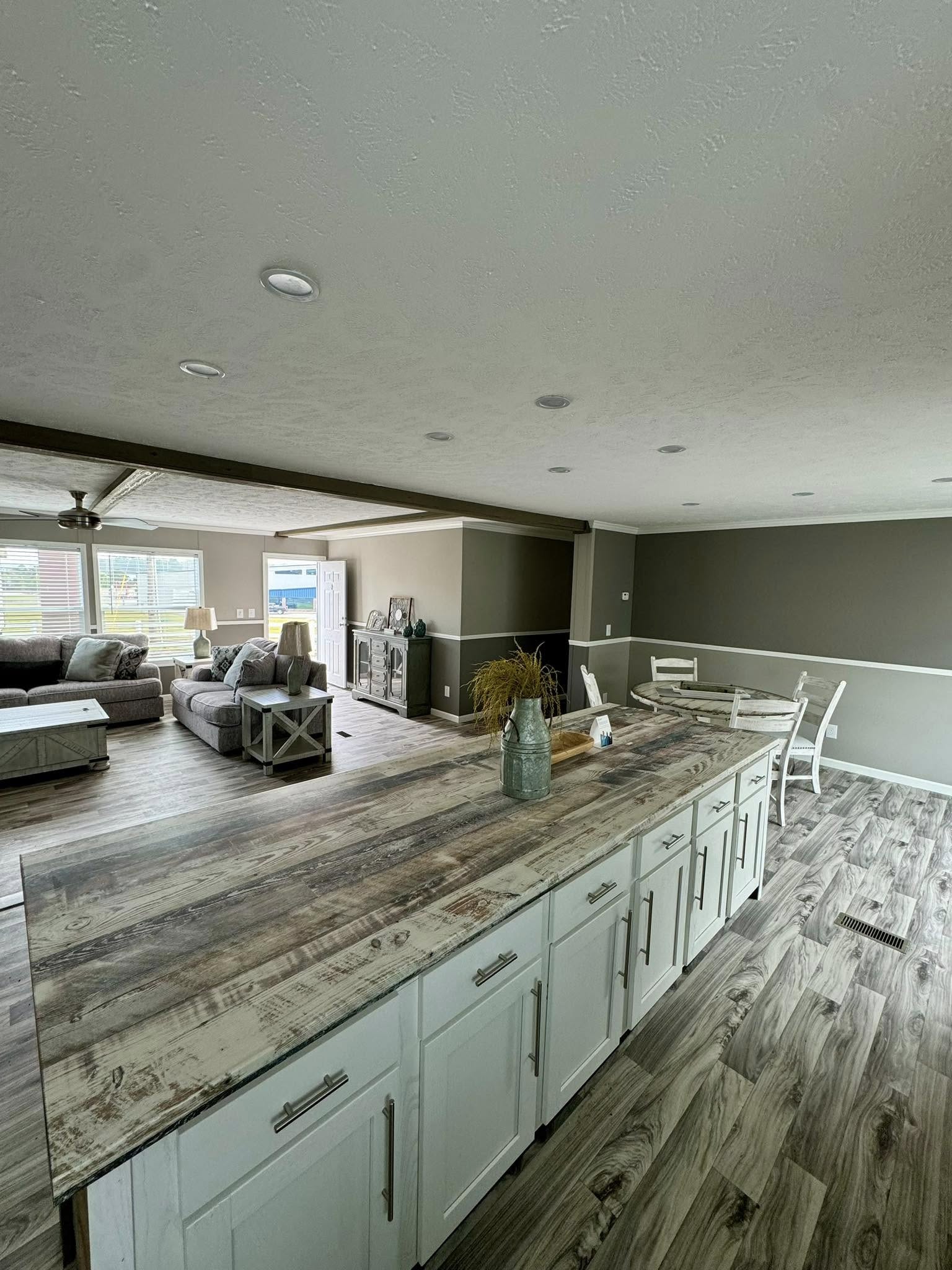 Spacious open-plan living room and kitchen with rustic wood floors, large island with decor, gray sofas, and soft natural lighting from windows.