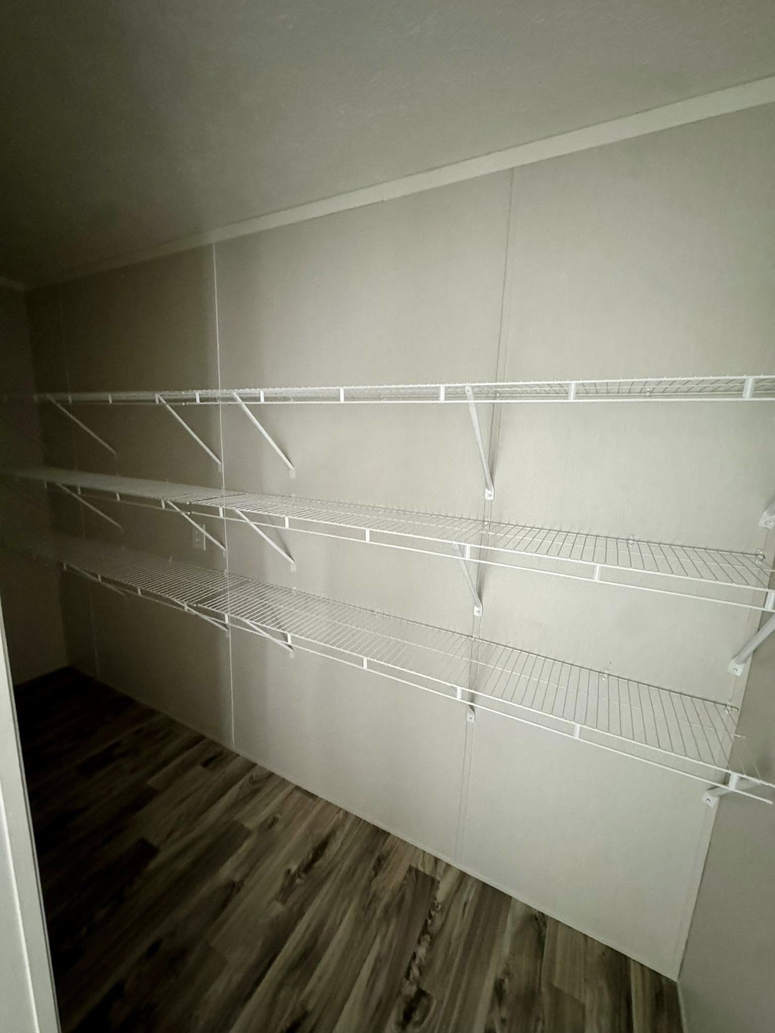 Empty closet with three white wire shelves mounted on a beige wall, over a dark hardwood floor. The space appears clean and organized.
