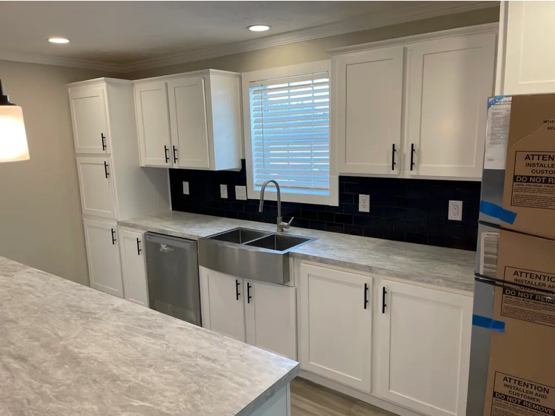 Modern kitchen with white cabinets, gray marbled countertops, and a stainless steel double sink. Black tile backsplash and a window with blinds above.