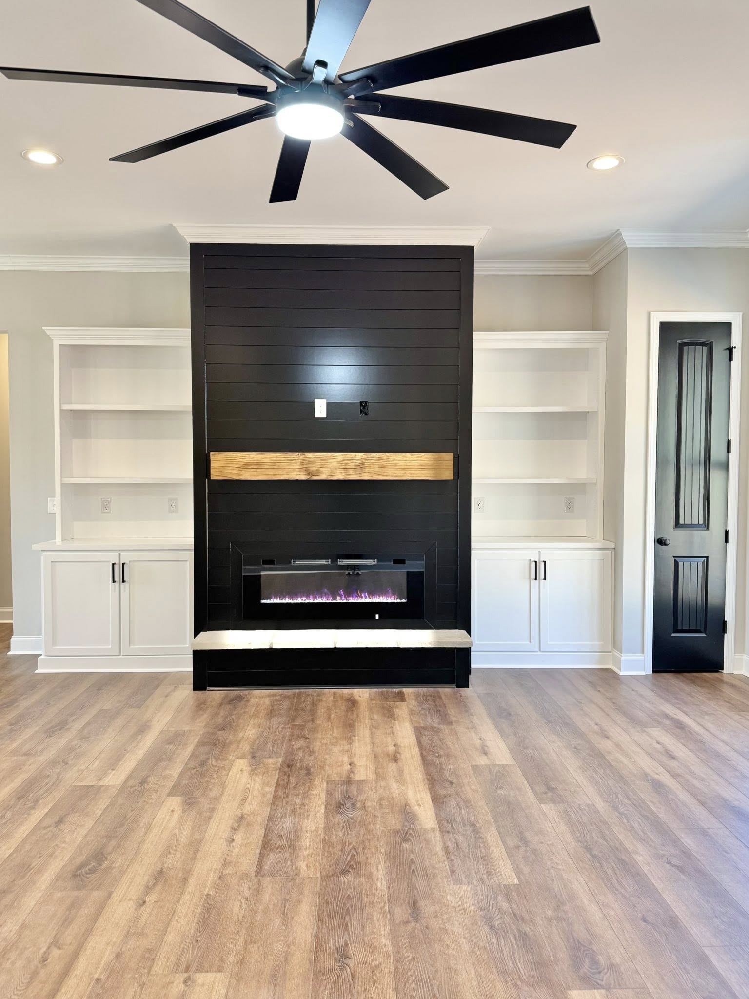 Spacious modern living room with a large ceiling fan, dark paneled fireplace, mounted light wood mantel, and built-in shelves. Light wood flooring adds warmth.