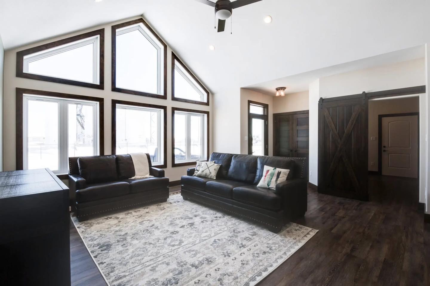 Bright living room with large, triangular windows, two black leather sofas, a patterned rug, dark wooden floor, and a sliding barn door. Airy and modern.