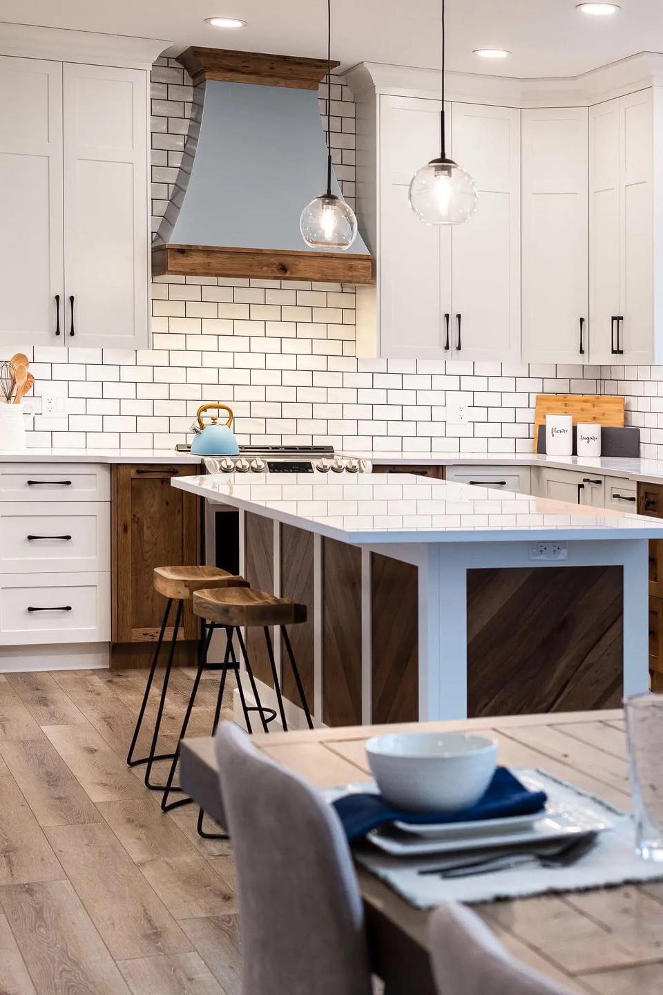 Modern kitchen with white cabinets, subway tile backsplash, and a wooden-accented range hood. Features island with stools, pendant lights, and cozy dining setup. Warm and inviting atmosphere.