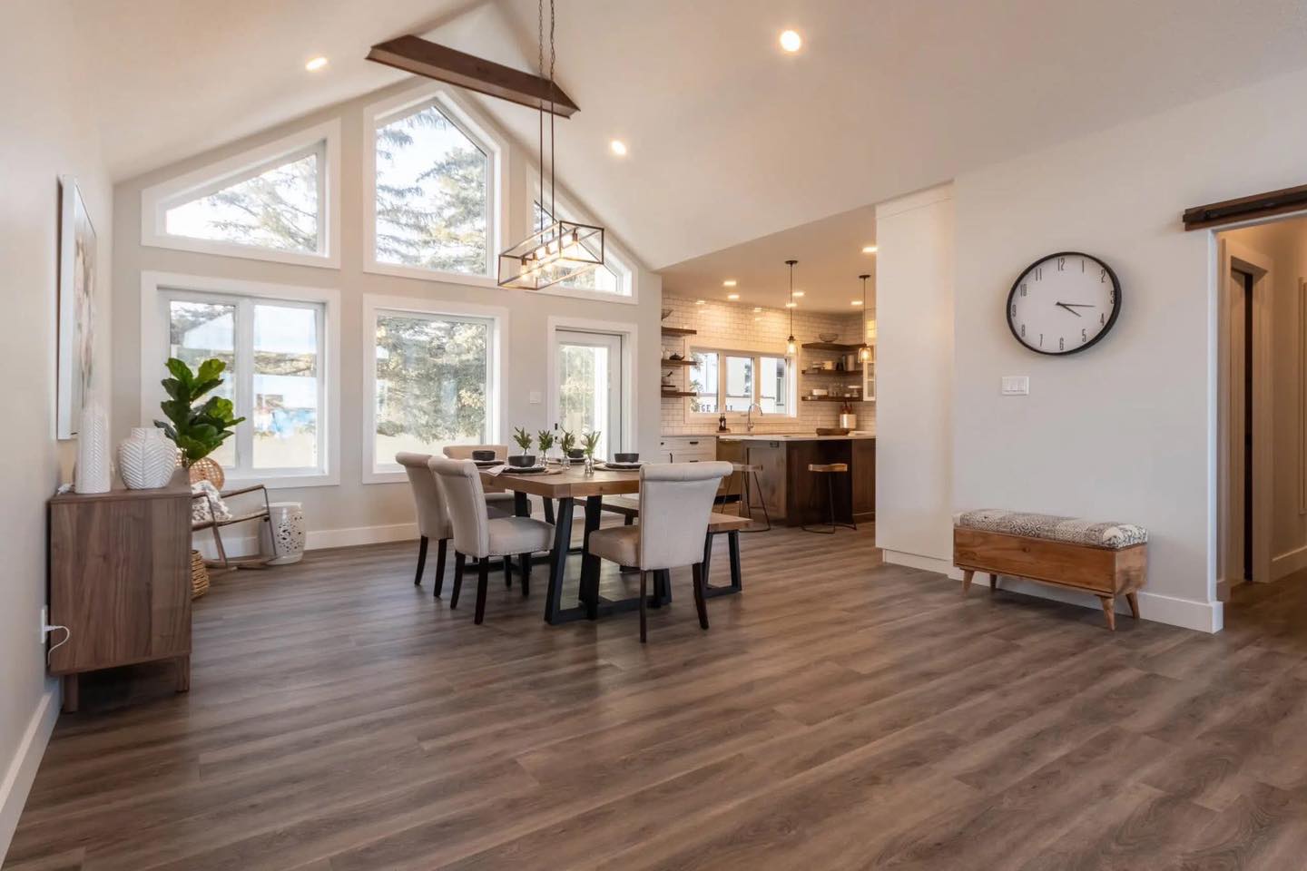 Spacious, modern dining room with high ceilings and large windows. Features a wooden table, six chairs, pendant lighting, and an open kitchen, exuding warmth.