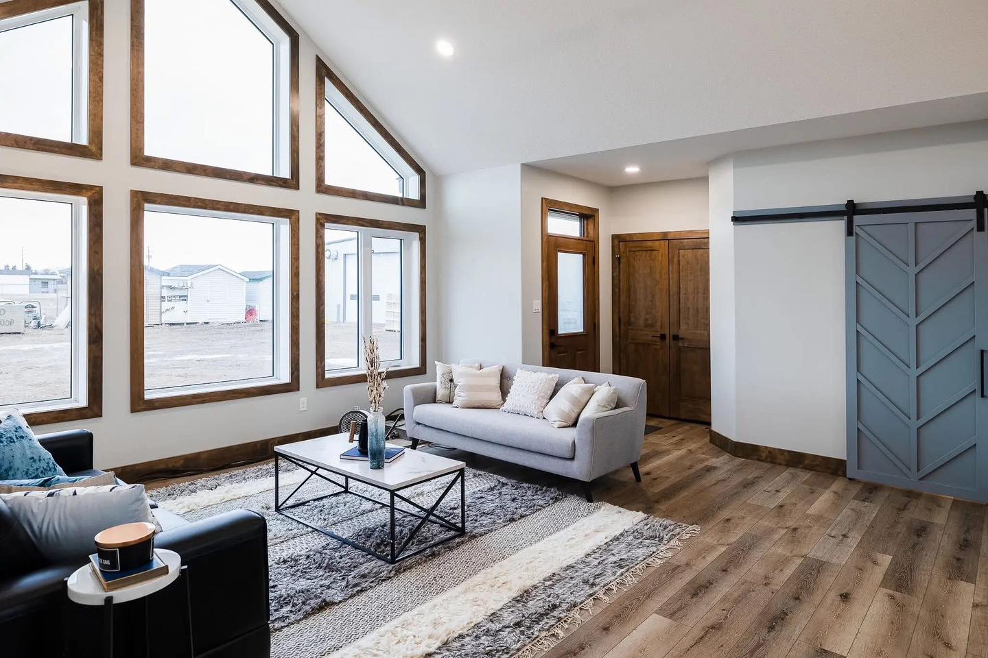 Spacious living room with large, triangular windows, light gray sofa, modern coffee table on a textured rug, wood flooring, and blue barn door. Cozy and bright.