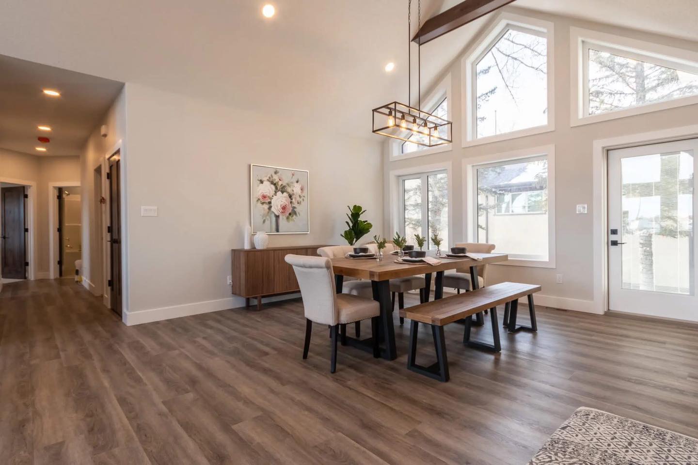 Bright dining area with high ceilings and wooden floors. Features a large table, modern light fixture, and three tall windows, creating an airy feel.