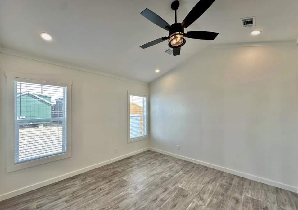 Bright, empty room with light wood flooring, white walls, and two large windows. A black ceiling fan hangs from a high ceiling, creating an airy feel.