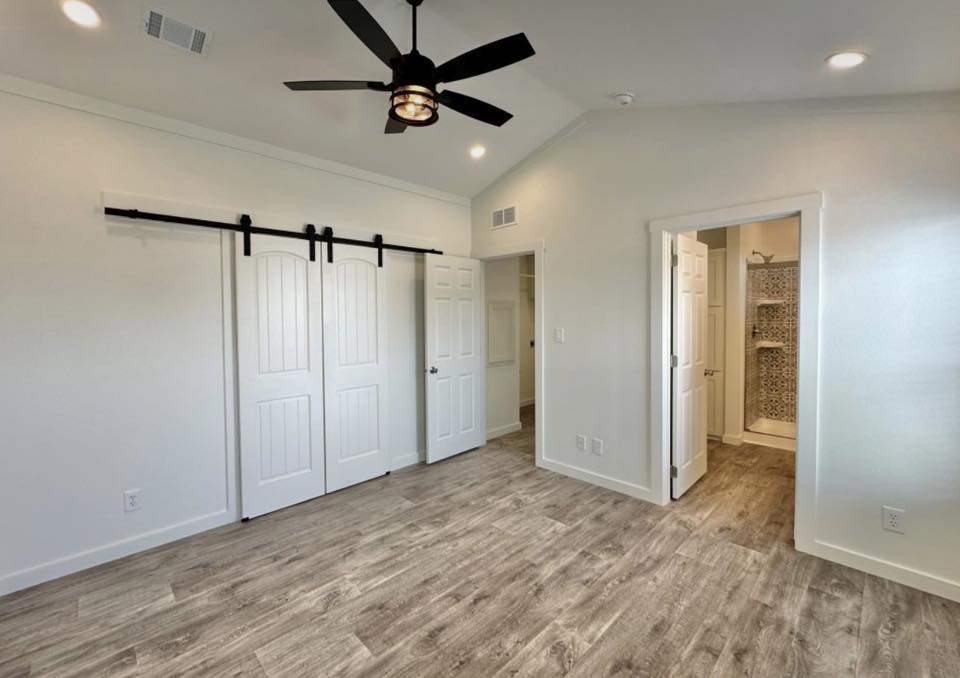 A modern room with light wood flooring, white walls, and a black ceiling fan. Features a barn door closet and open door to a bathroom with a tiled shower.