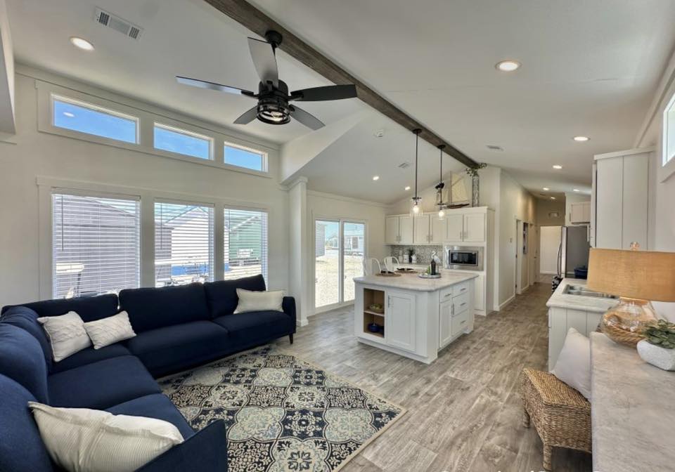 Spacious living room with a navy L-shaped sofa, patterned rug, and ceiling fan. Open kitchen with white cabinets and island. Bright, airy ambiance.