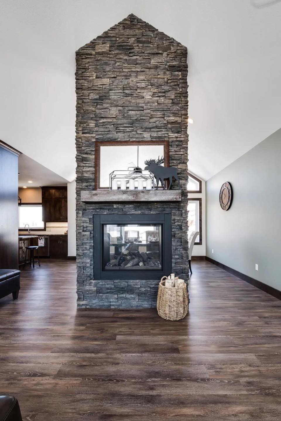 Modern living room with a tall stone fireplace, wooden floors, and vaulted ceiling. Decor includes a clock on the wall and a wicker basket near the hearth.