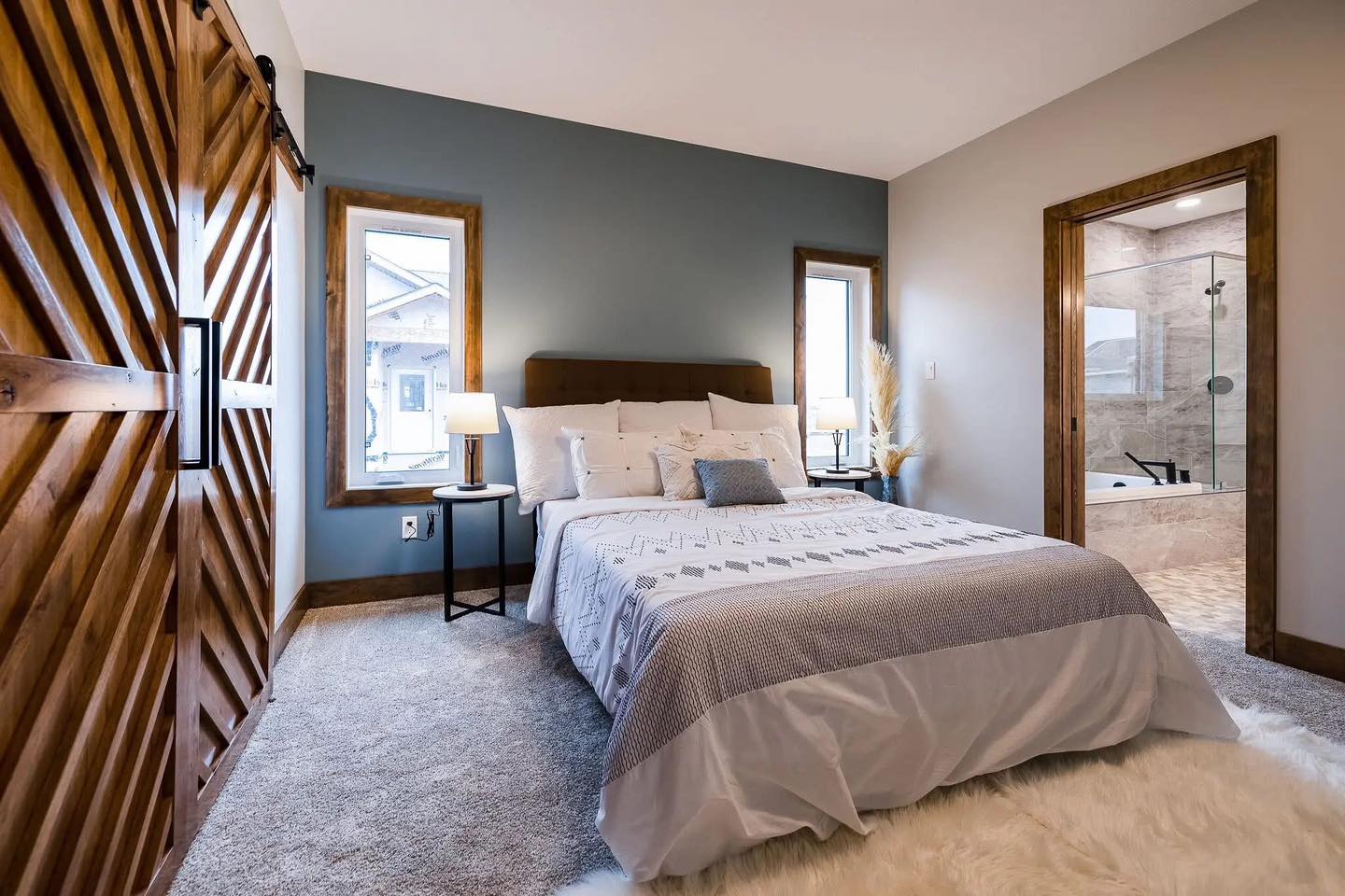 A cozy bedroom with a blue accent wall, a neatly made bed with white and gray bedding, two windows, a wooden sliding door, and an en-suite bathroom.