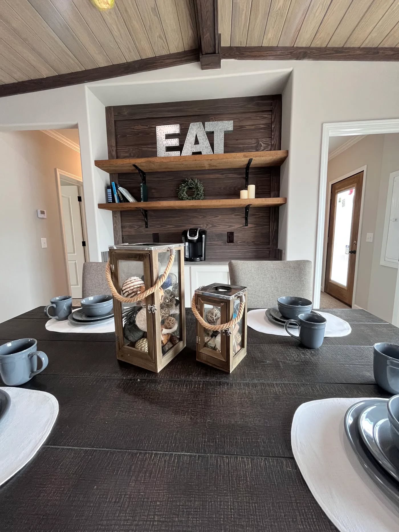 Dining room with a dark wooden table set for six, featuring gray dishes and cups. Two decorative lanterns with seashells are centered. A wall displays shelves, a coffee machine, and large 'EAT' letters, creating a cozy, rustic ambiance.