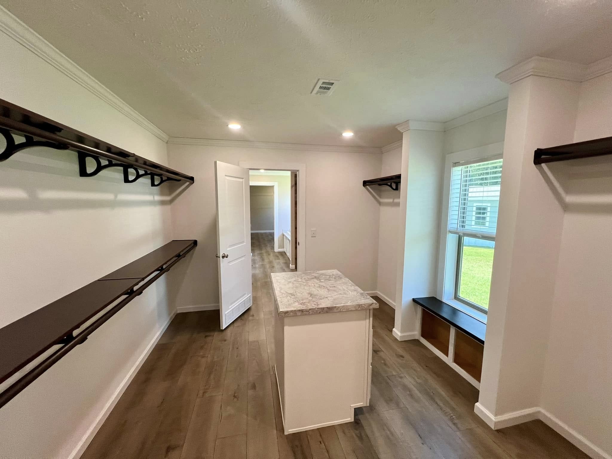 Spacious walk-in closet with wooden shelves, a central marble-topped island, and a large window seat. The room has a bright and modern feel.