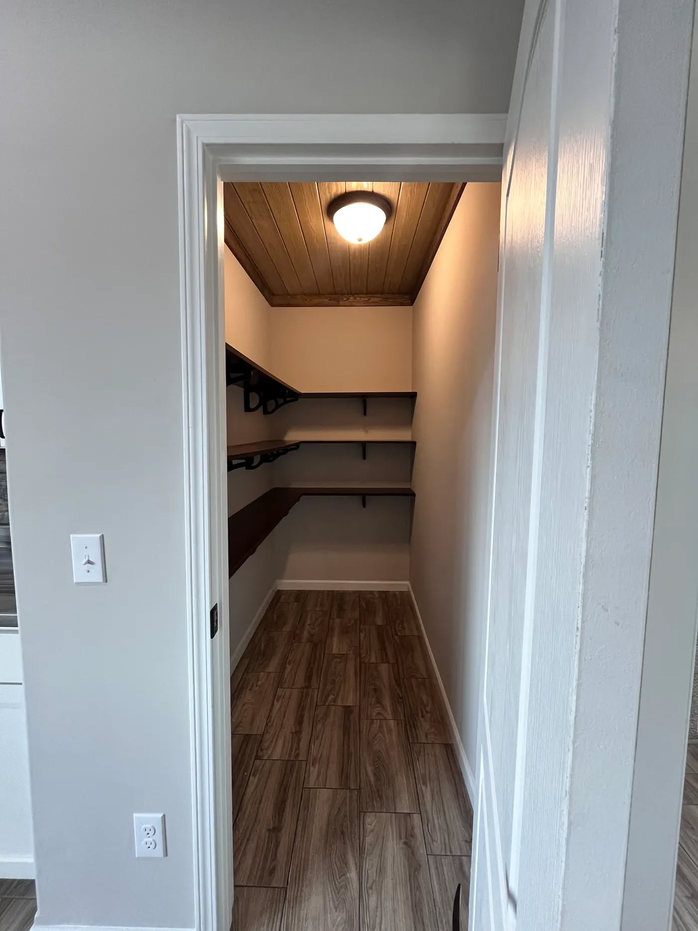 Small walk-in closet with wooden flooring and ceiling, lit by a round ceiling light. Dark wooden shelves line the left wall, creating a warm, cozy atmosphere.