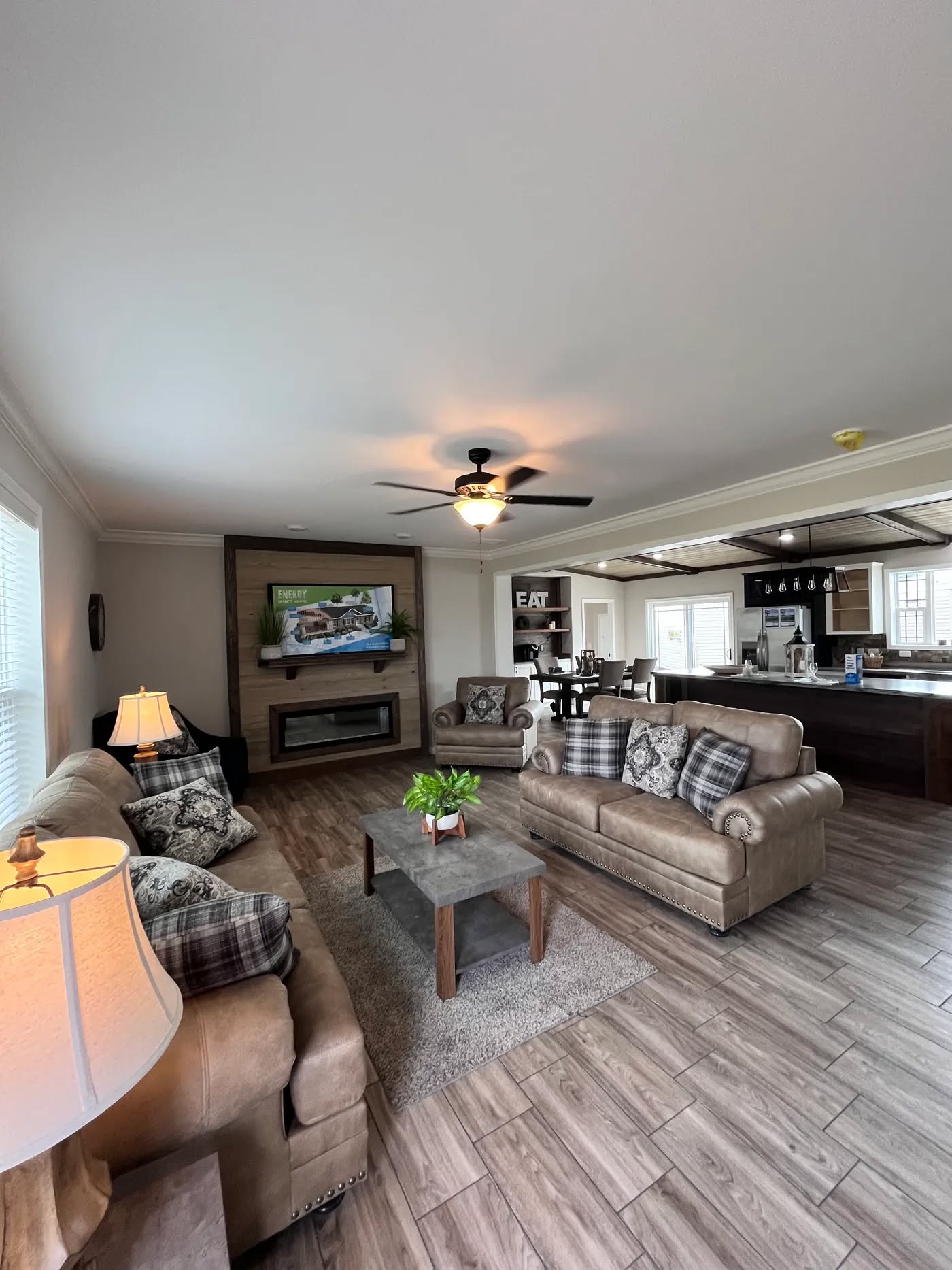 Spacious living room with brown sofas, plaid cushions, and a wooden coffee table. Warm lighting, ceiling fan, open kitchen and dining area in view.