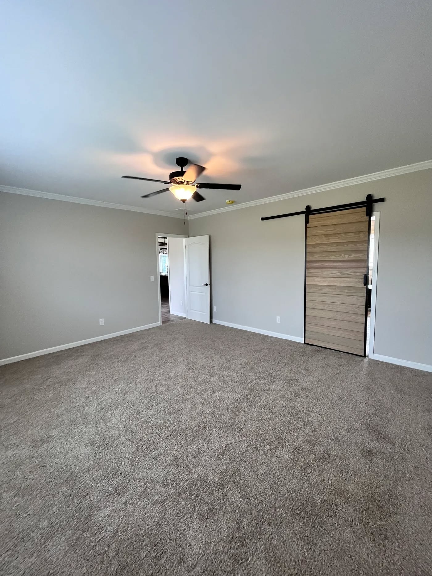 Spacious room with beige carpet, a ceiling fan with light, gray walls, and a sliding wooden barn door. Open door reveals an adjacent room. Cozy and modern ambiance.