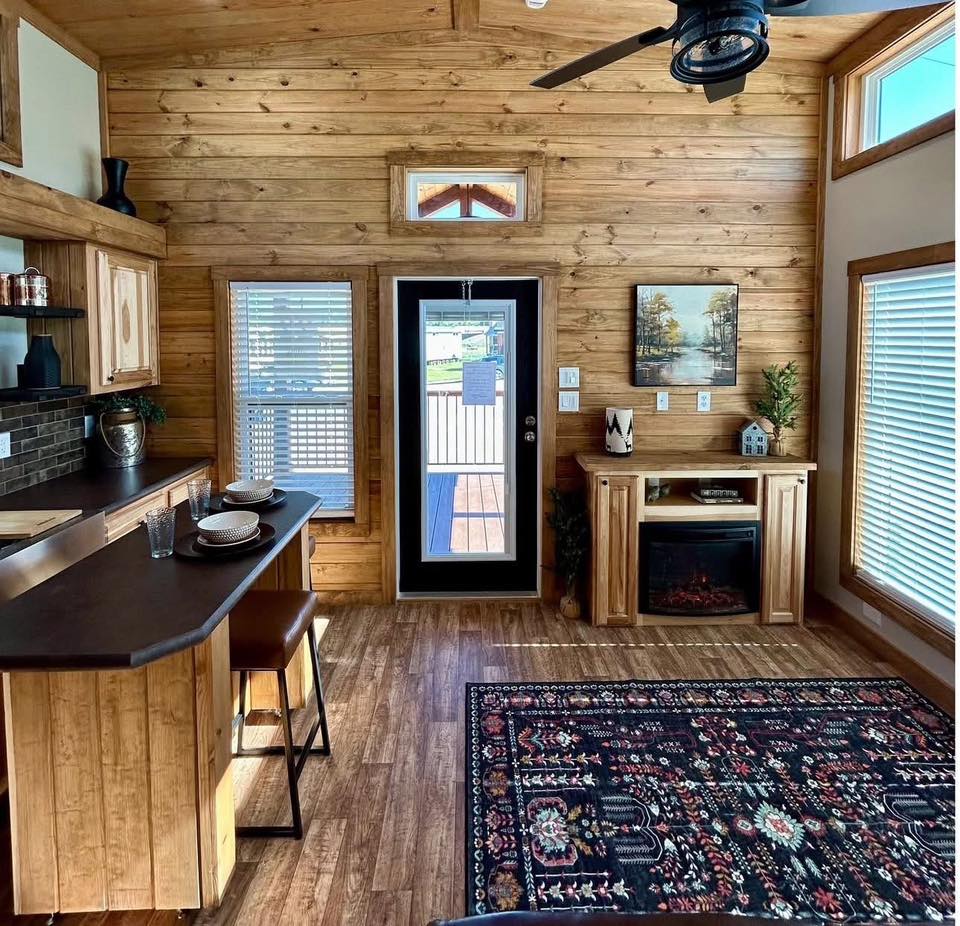 Warm, cozy cabin interior with wood-paneled walls, a black countertop with stools, and a vibrant patterned rug. A door leads to a sunlit porch.