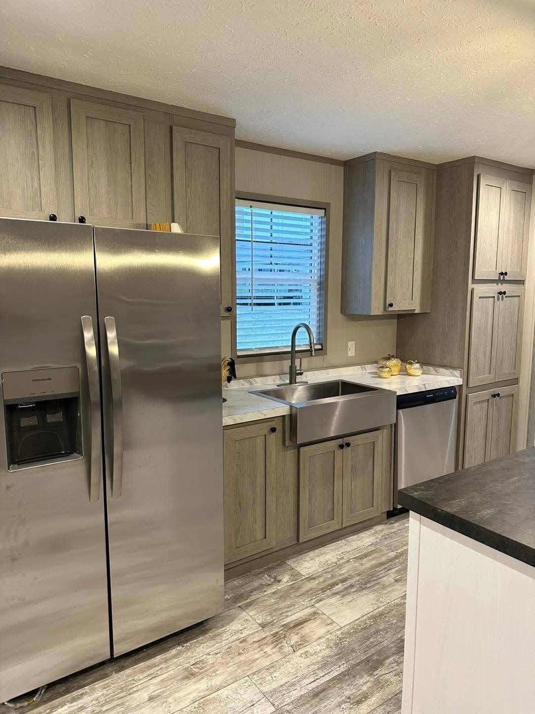 Modern kitchen with gray wood cabinets, a stainless steel fridge, farmhouse sink, and dishwasher. Warm lighting and wood-patterned floor create a cozy feel.