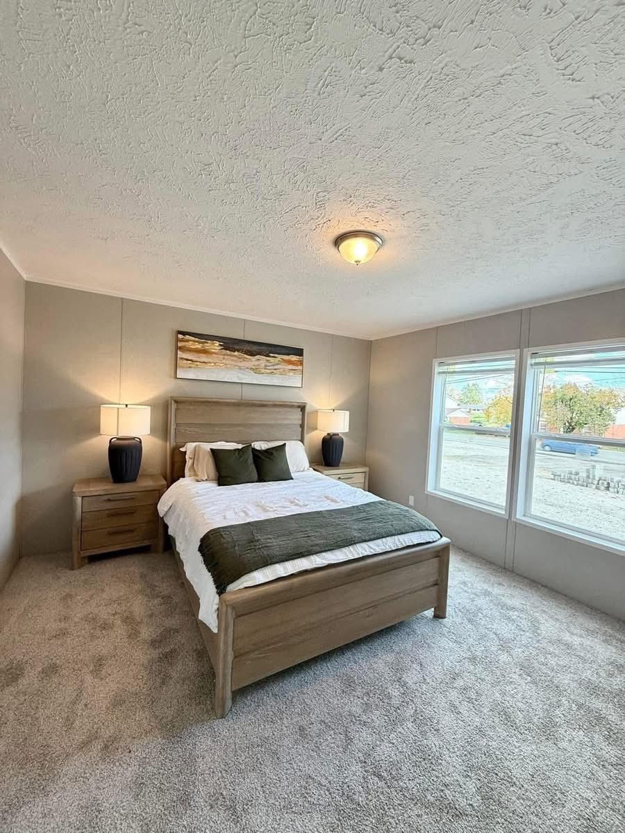 A cozy bedroom with a wooden bed featuring white bedding and green pillows, flanked by two nightstands with black lamps. A landscape painting hangs above. Bright natural light streams through large windows, highlighting the soft carpet and neutral wall tones, creating a calm atmosphere.