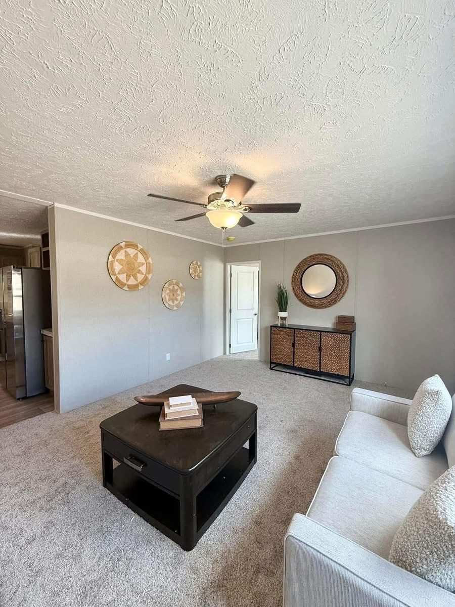 A cozy living room with neutral tones featuring a beige sofa, a dark square coffee table, a textured rug, circular wall art, and a ceiling fan adding warmth.