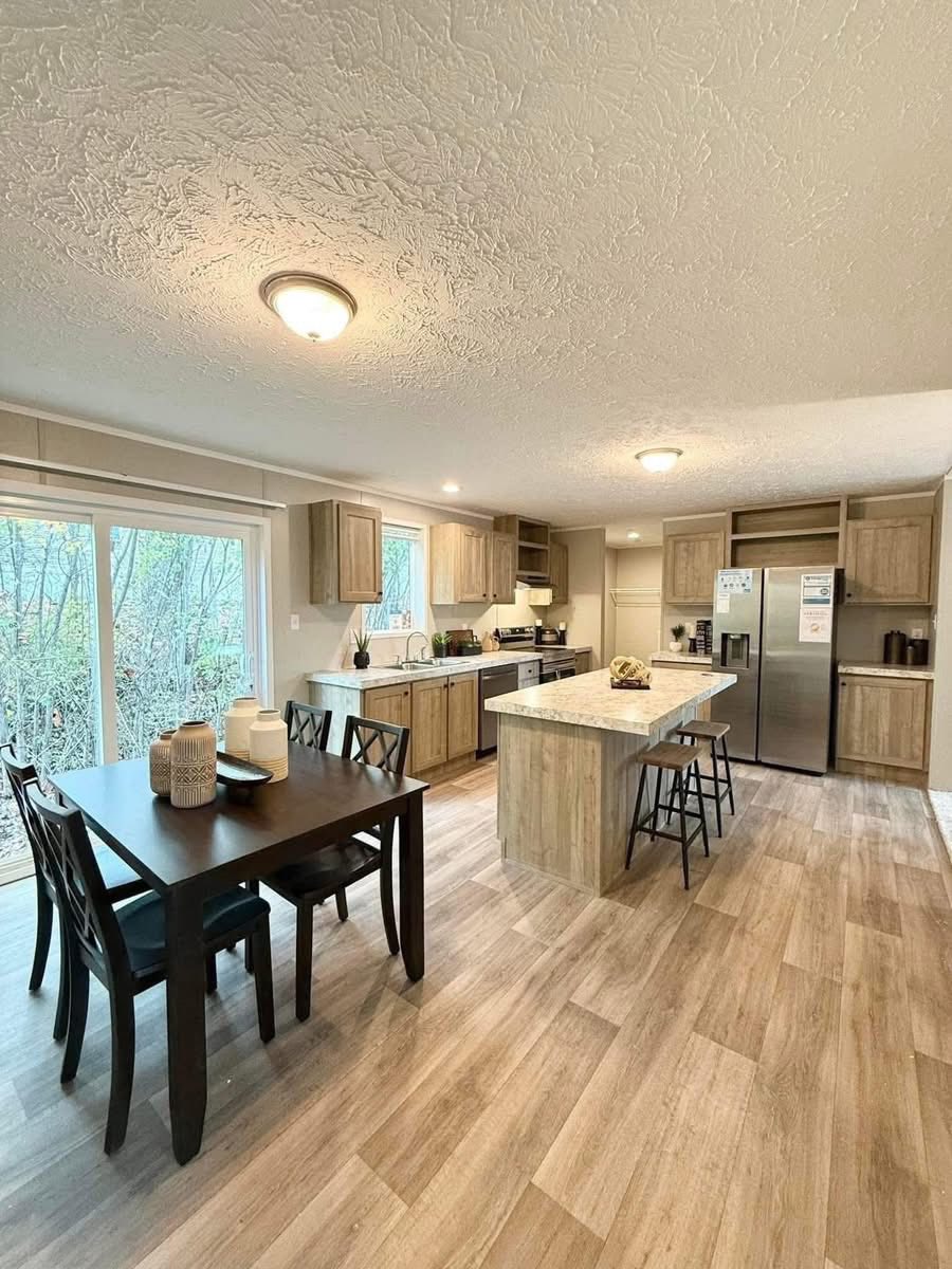 Modern kitchen with light wood cabinets, stainless steel appliances, and an island with bar stools. A dining table with chairs sits near large windows, creating a bright, inviting space.