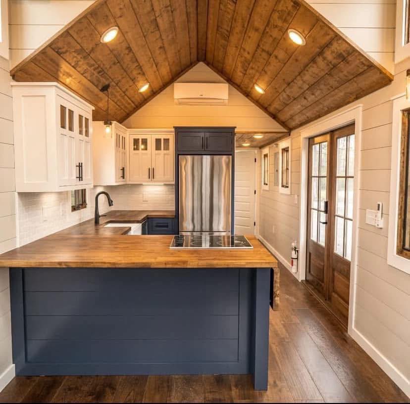 Cozy kitchen with rustic wood ceiling, navy island, and butcher block counters. Modern appliances, white cabinets, and large wooden door. Warm, inviting feel.