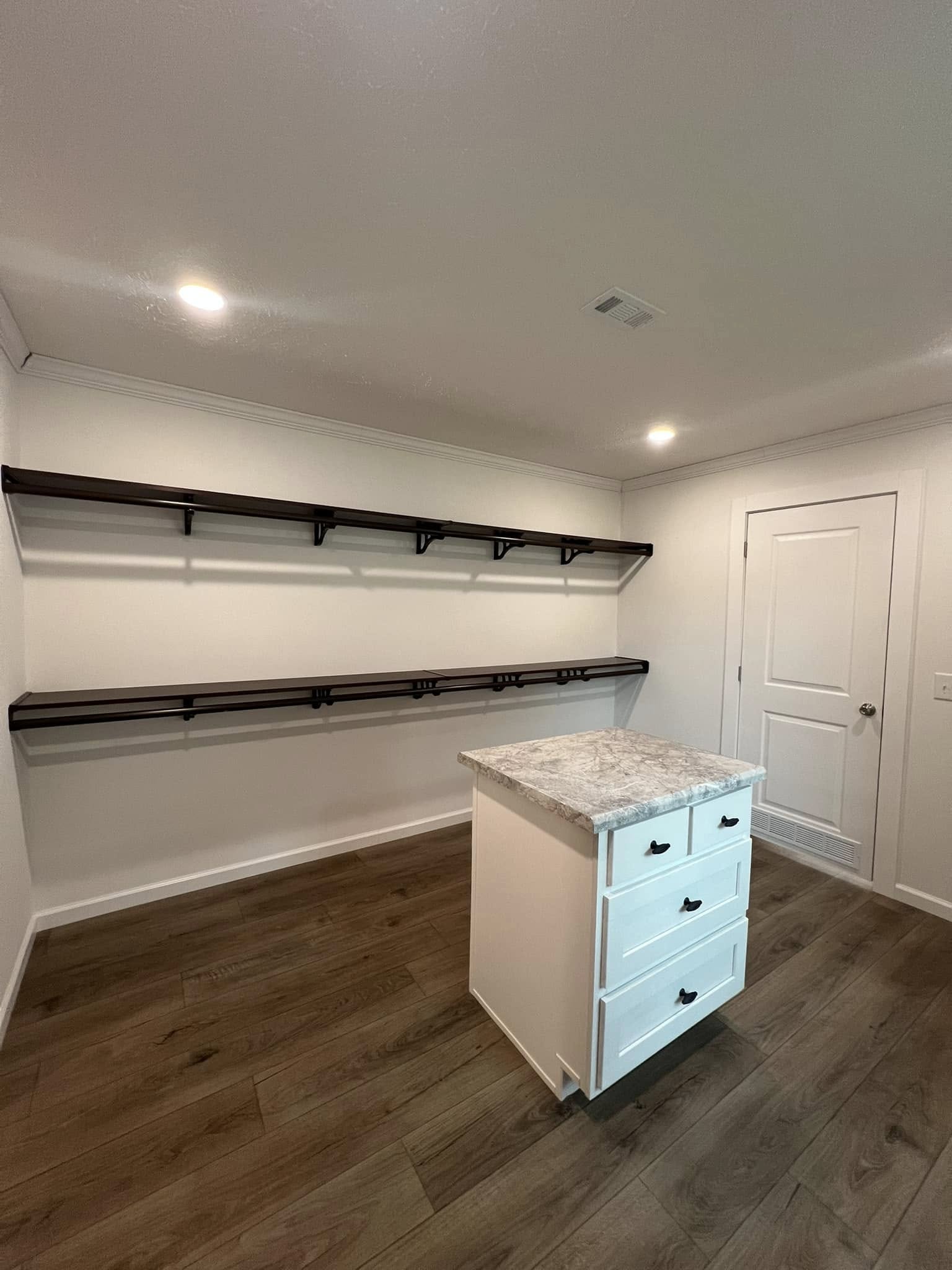 Minimalist closet with white walls and dark wood flooring. Features an island with drawers and granite countertop. Floating shelves line the walls, lit by recessed lighting.