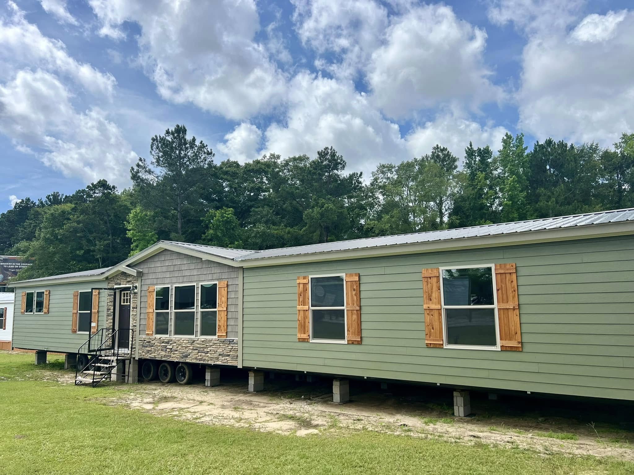 A mobile home with stone accents and wooden shutters on windows. It sits on wheels with a small black staircase leading to the entrance, set against a backdrop of trees under a cloudy sky.