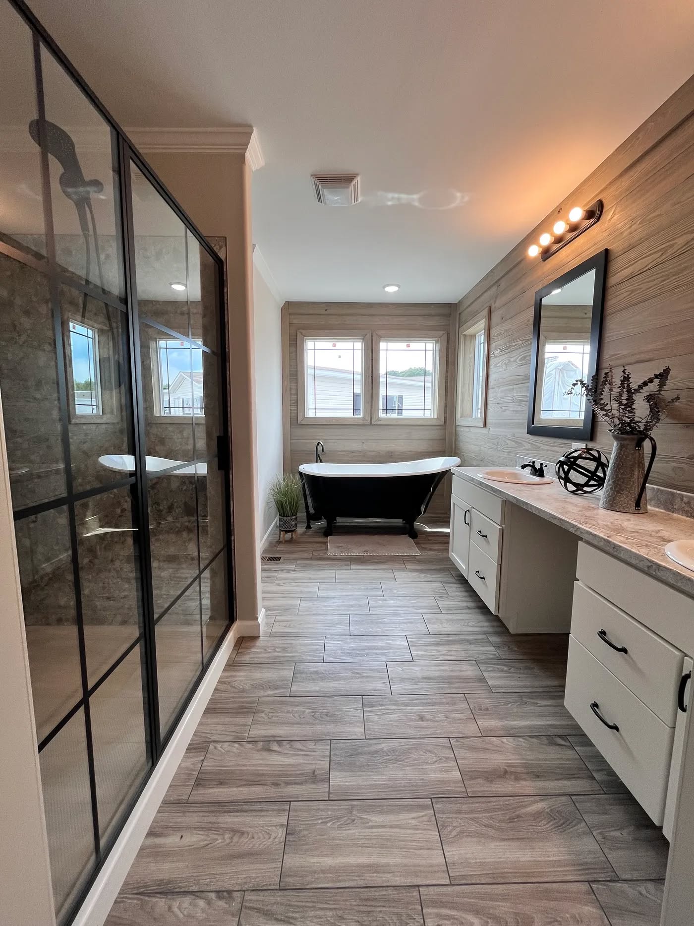 Modern bathroom with a wood-patterned tile floor, glass shower, freestanding black tub under windows, and vanity with a mirror. Warm lighting and a cozy feel.