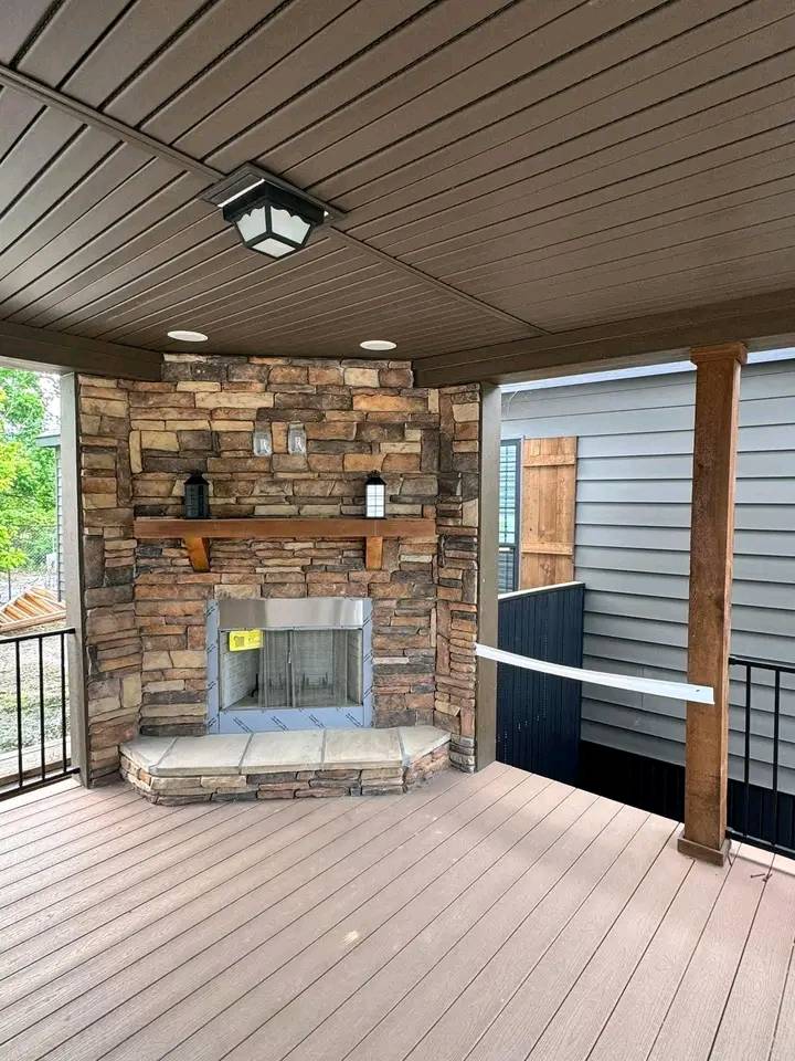 Covered patio with a wooden ceiling and brown decking. A stone fireplace, with a wooden mantel holding two decorative items, adds coziness.