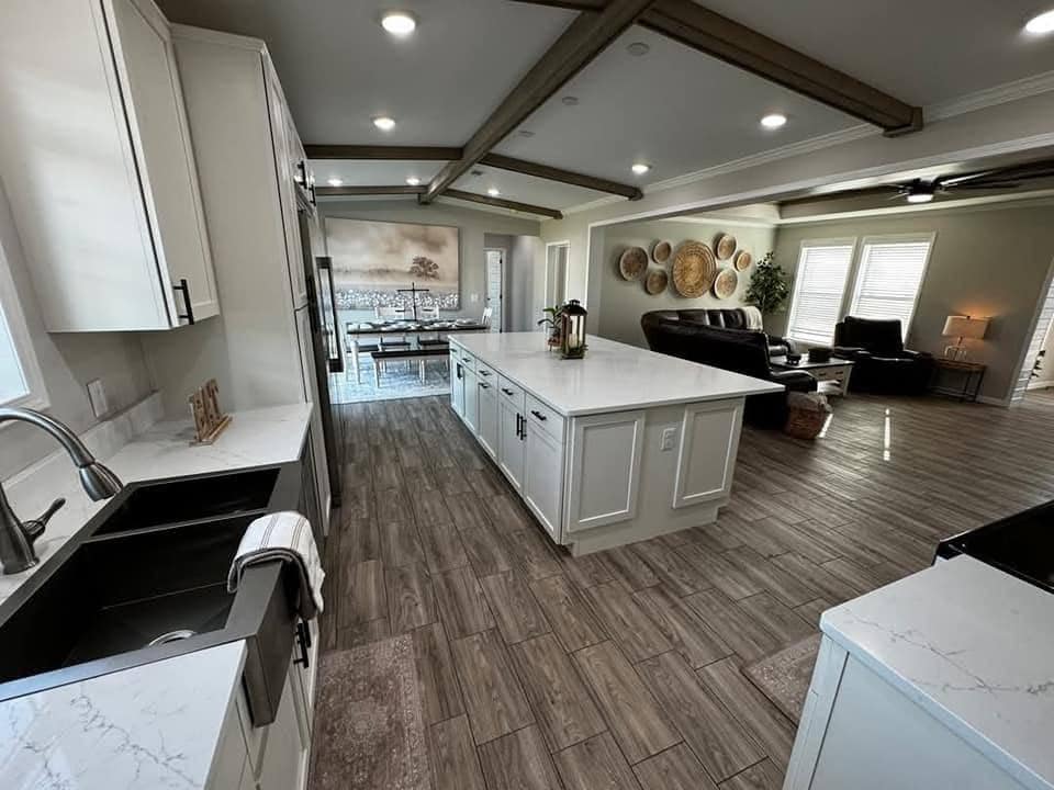 Spacious kitchen with white cabinets, marble countertops, and center island. Open living area features wooden floors, beam ceiling, and cozy seating.