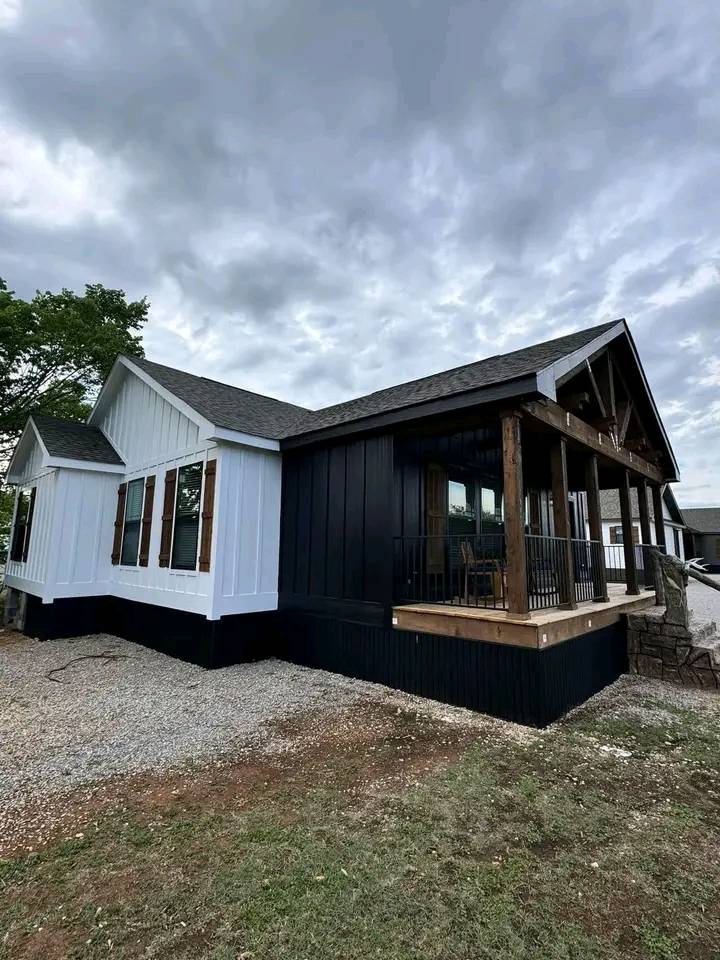 Modern house with white and black siding, featuring large windows and a porch with wooden beams. Overcast sky enhances a cozy, rustic vibe.
